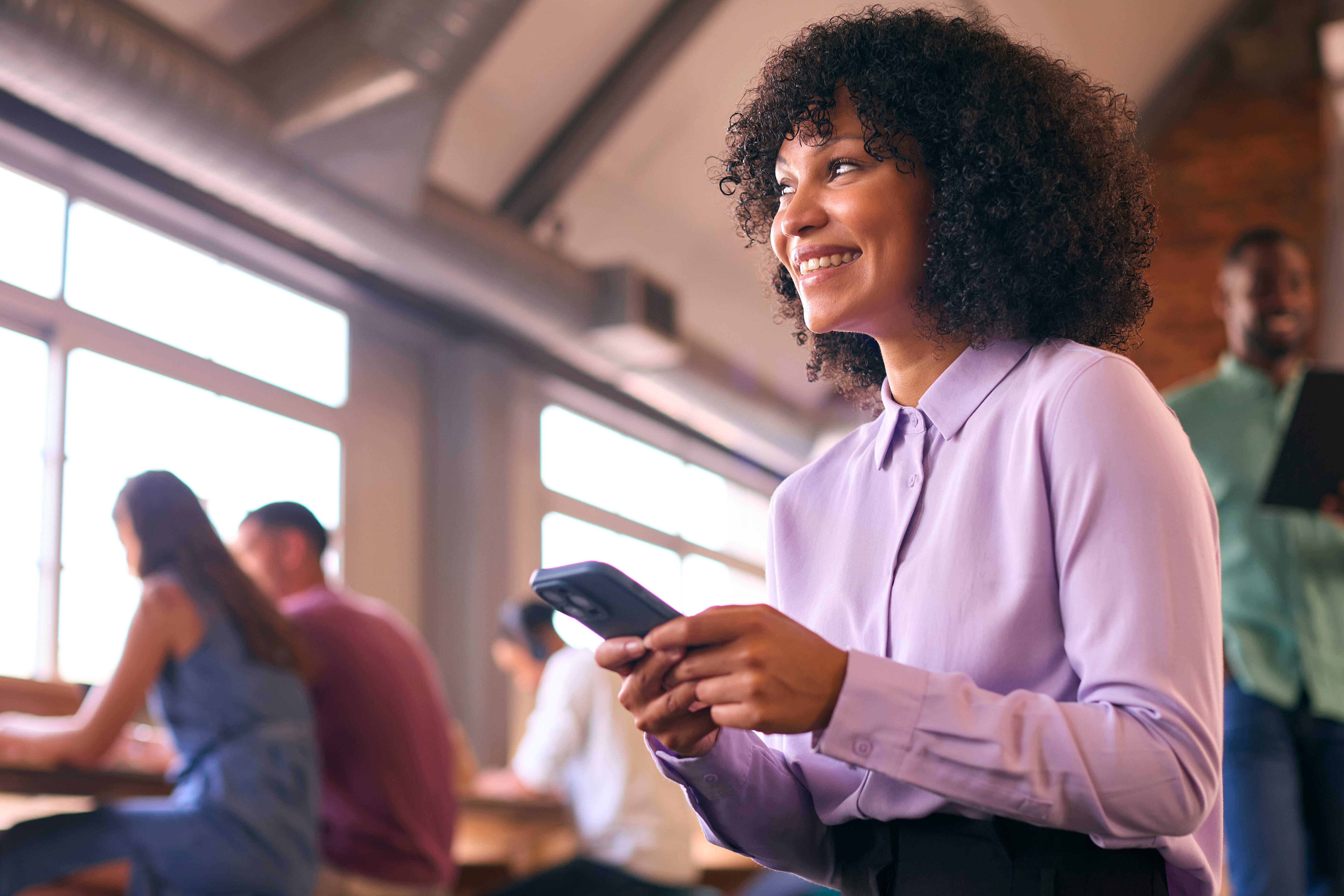 smiling-businesswoman-with-mobile-phone-working-open-plan-office-with-colleagues-background (1)